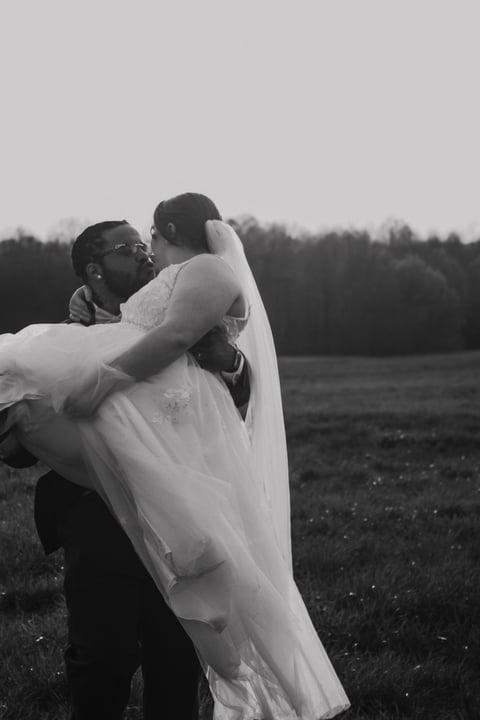Groom carrying bride in white wedding dress in open field, black and white photography
