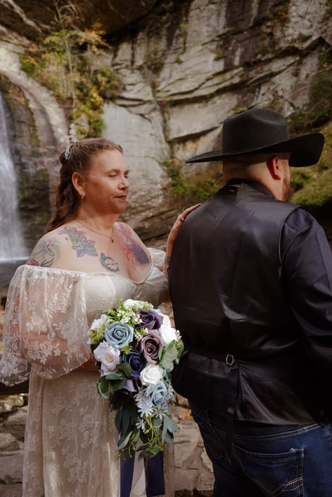 Bride in lace dress holding purple and white flowers standing with groom in black cowboy hat and jacket near a waterfall