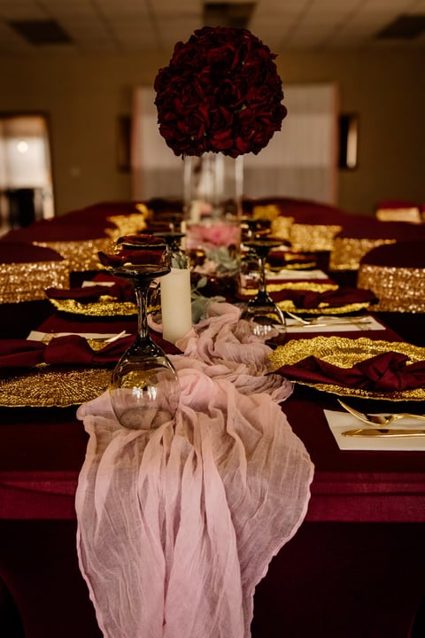 Elegant formal dining table with deep red roses, burgundy linens, gold napkins, and pink tulle runner