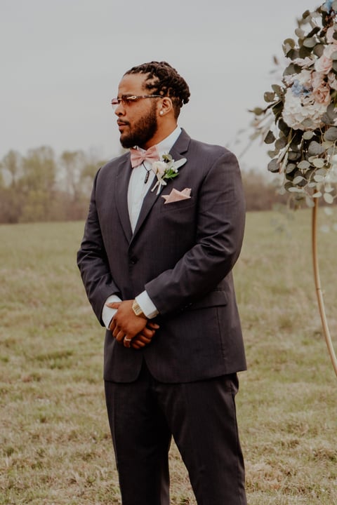 Groom in dark charcoal suit and glasses standing in field next to floral wedding arch, looking to the side