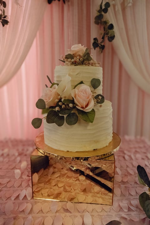 Three-tier white wedding cake with pink and cream roses, eucalyptus leaves, on gold stand