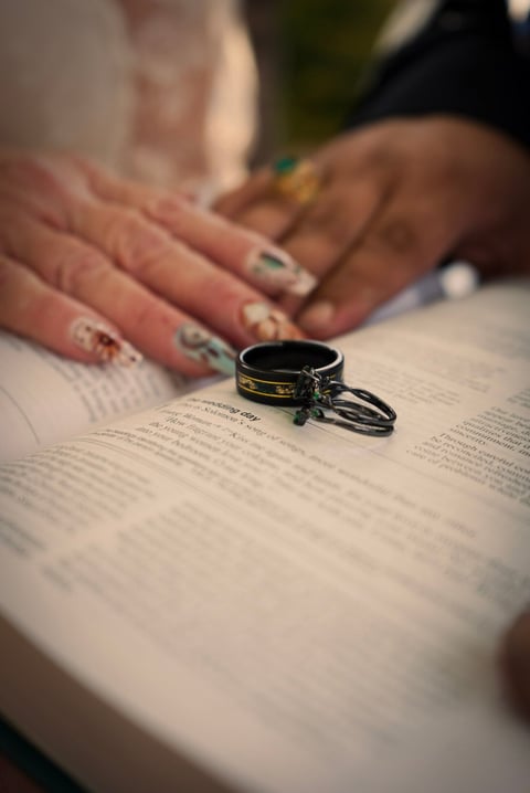 Wedding rings displayed on an open book with couples hands showing engagement rings in background