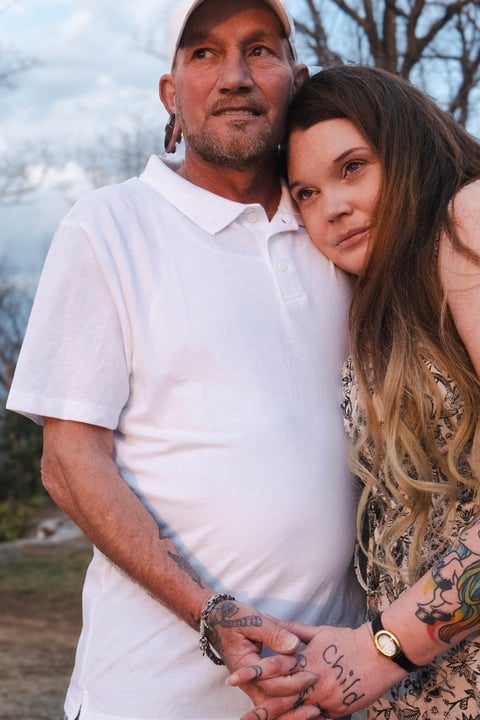 A man and woman posing together outdoors, both smiling at the camera with bare trees visible in the background