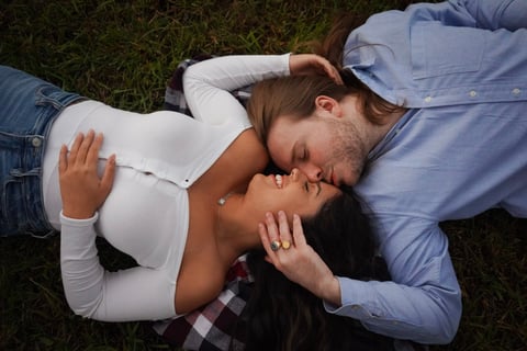 A couple lying on grass, kissing while looking down at the camera in an overhead shot
