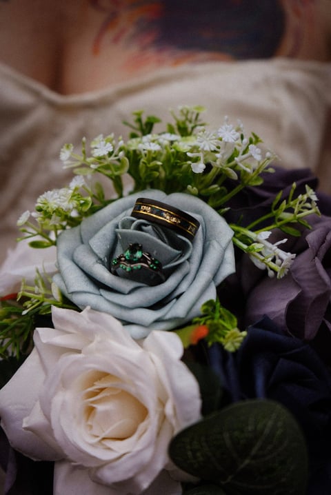 Wedding rings displayed on a light blue fabric rose, surrounded by white flowers and greenery in a bridal bouquet