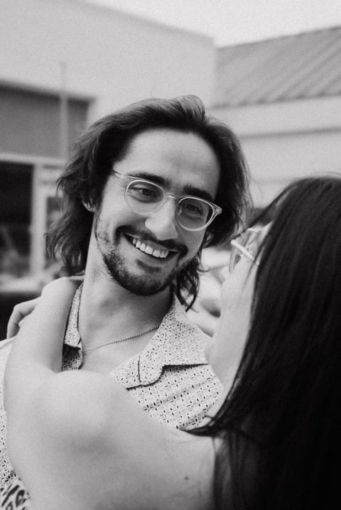 Man with long dark hair and glasses smiling at camera in black and white photo outdoors